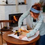 A woman is cutting paper on a table, focused on her task with scissors in hand and sheets of paper spread out